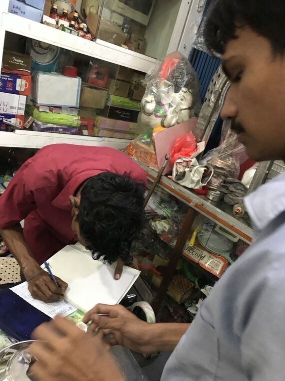 店の中で男性2人が対応している。ひとりが紙に手書きで伝票を書いていて、背後の棚にガネーシャ像が置かれている