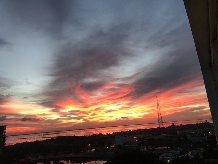 ラグーンの向こうに広がる真紅の夕焼け。厚い雲の下が赤とオレンジに染まり、右手に鉄骨の通信塔と街並みのシルエットが見える