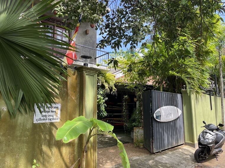 黄土色の塀とDON'T DUMP YOUR GARBAGE HEREのシンハラ語併記の看板。灰色のゲートにflowのロゴ入りサーフボード。奥の敷地にサーフボードが並ぶ