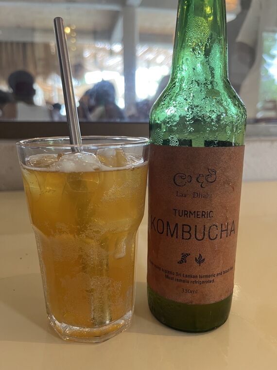 緑の瓶に貼られたクラフト紙のラベル。シンハラ文字のロゴとLaa Dhahi、TURMERIC KOMBUCHA、330mlの表記。左に氷を入れたグラスに注がれたオレンジがかった黄色の液体
