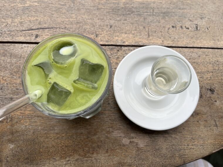 使い込まれた木のテーブルに置かれた明るい黄緑色のアイス抹茶ラテを真上から見たところ。大きめの角氷とガラスのストロー。右にソーサー付きのチェイサーの水