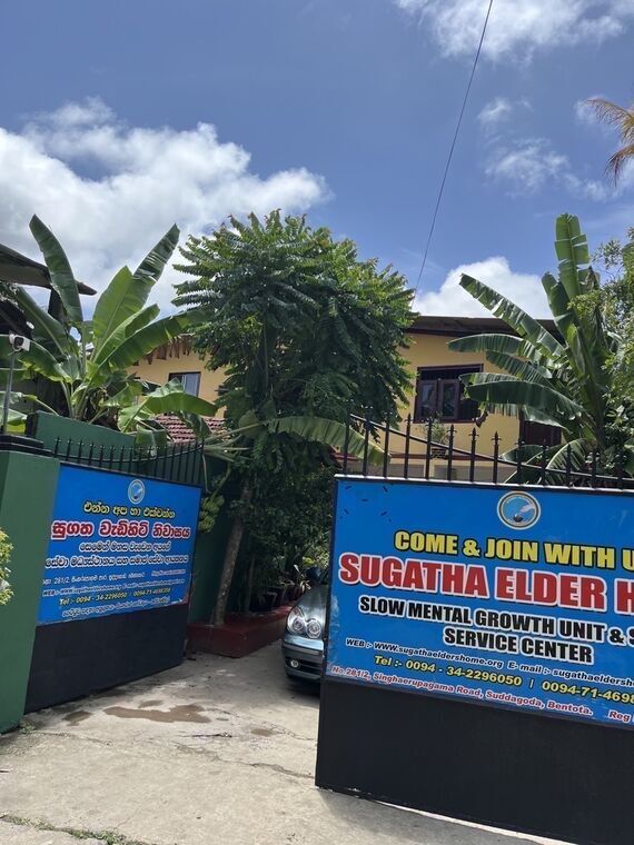 Sugatha Elders' Homeの正面ゲート。青い看板にSUGATHA ELDER HOME、SLOW MENTAL GROWTH UNIT &amp; SOCIAL SERVICE CENTERの表記。奥に黄色い2階建ての建物とバナナの木