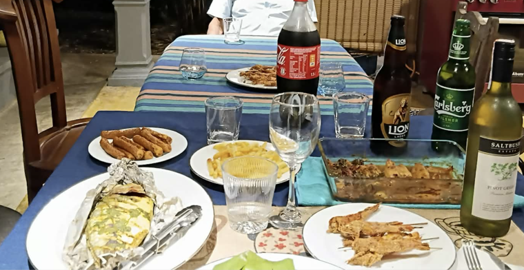 お祝いの食卓。ホイル包み焼きの魚、揚げ物の串、フライドポテト、耐熱ガラス皿の料理と、Lion Lager、Carlsberg、白ワインのボトルが並ぶ