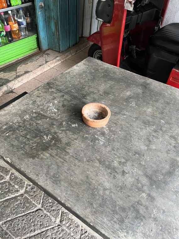 店先のコンクリートのテーブルに置かれた素焼きの小鉢。内側が黒く煤けて灰皿として使われている。奥に赤いトゥクトゥク