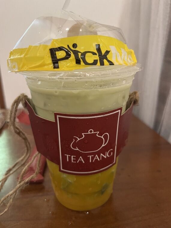 Amba Matchaのカップ。透明カップに黄緑色の抹茶ラテ、底に黄色いマンゴーの層。蓋に黄色いPickMeのシール、赤いTEA TANGのスリーブ