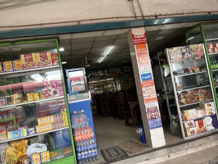 Cafe 'N' Chillの入口。左右のガラスケースにビスケットやケーキ、中央の柱に通信会社のステッカー、奥に木のテーブル席が並ぶ食堂スペース