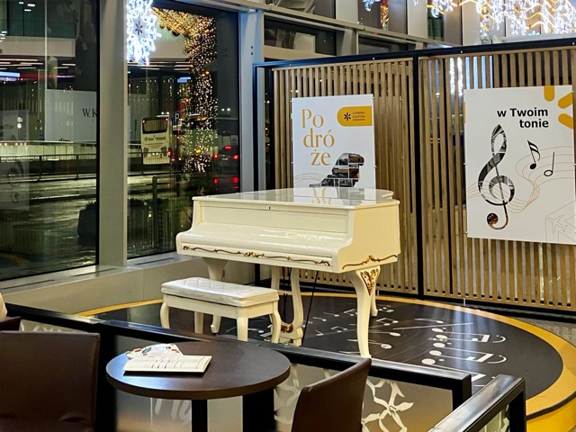 ワルシャワ空港ターミナルに設置された白いグランドピアノ。壁には空港ロゴ入りのポスターと大きなト音記号の装飾がある