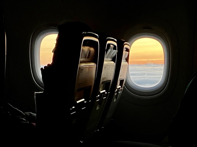 オーストリア航空の機内から望む夕暮れ。窓の外に広がるオレンジ色の雲海と、機内の座席シルエット