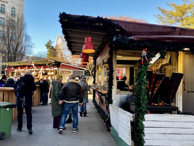ブダペストの街角に並ぶクリスマスマーケットの屋台。12月末の青空と屋台の装飾、奥に見える観覧車