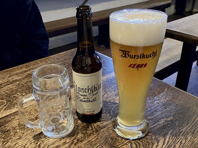 木製のテーブルに置かれたBrauerei Jacobの瓶ビールとジョッキ、ロゴ入りグラスの白ビール