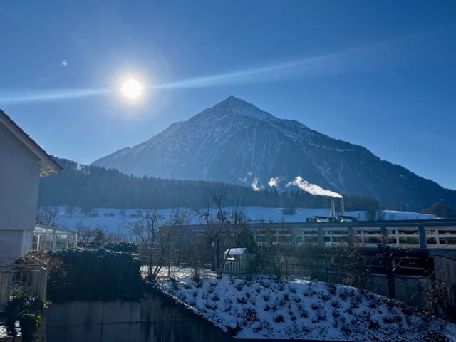 シュピーツの高台から見上げる、太陽の光に照らされた冬のニーゼン山と煙突から煙がのぼる風景
