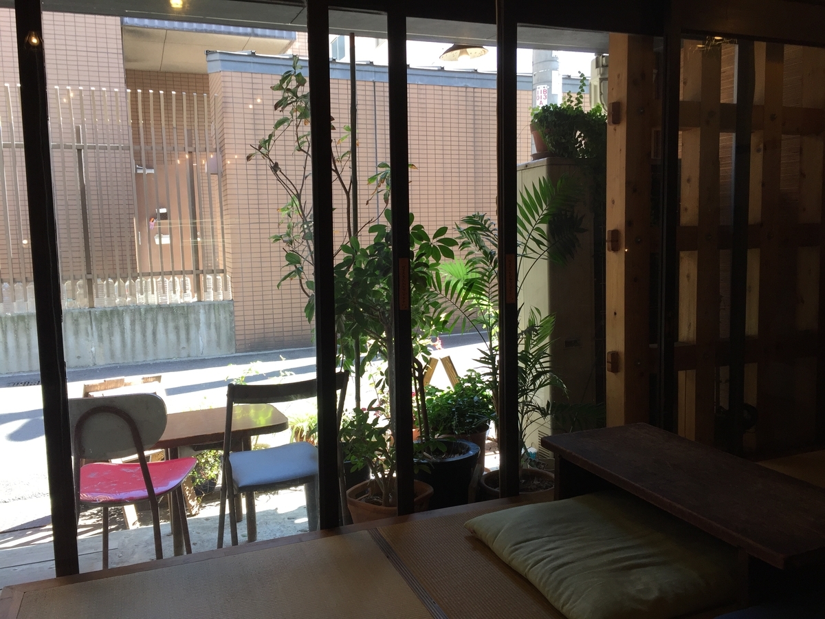 店内から入口のガラス戸越しの景色。入り口の鉢植えと喫煙席らしき灰皿の置かれたテーブルと椅子が一脚