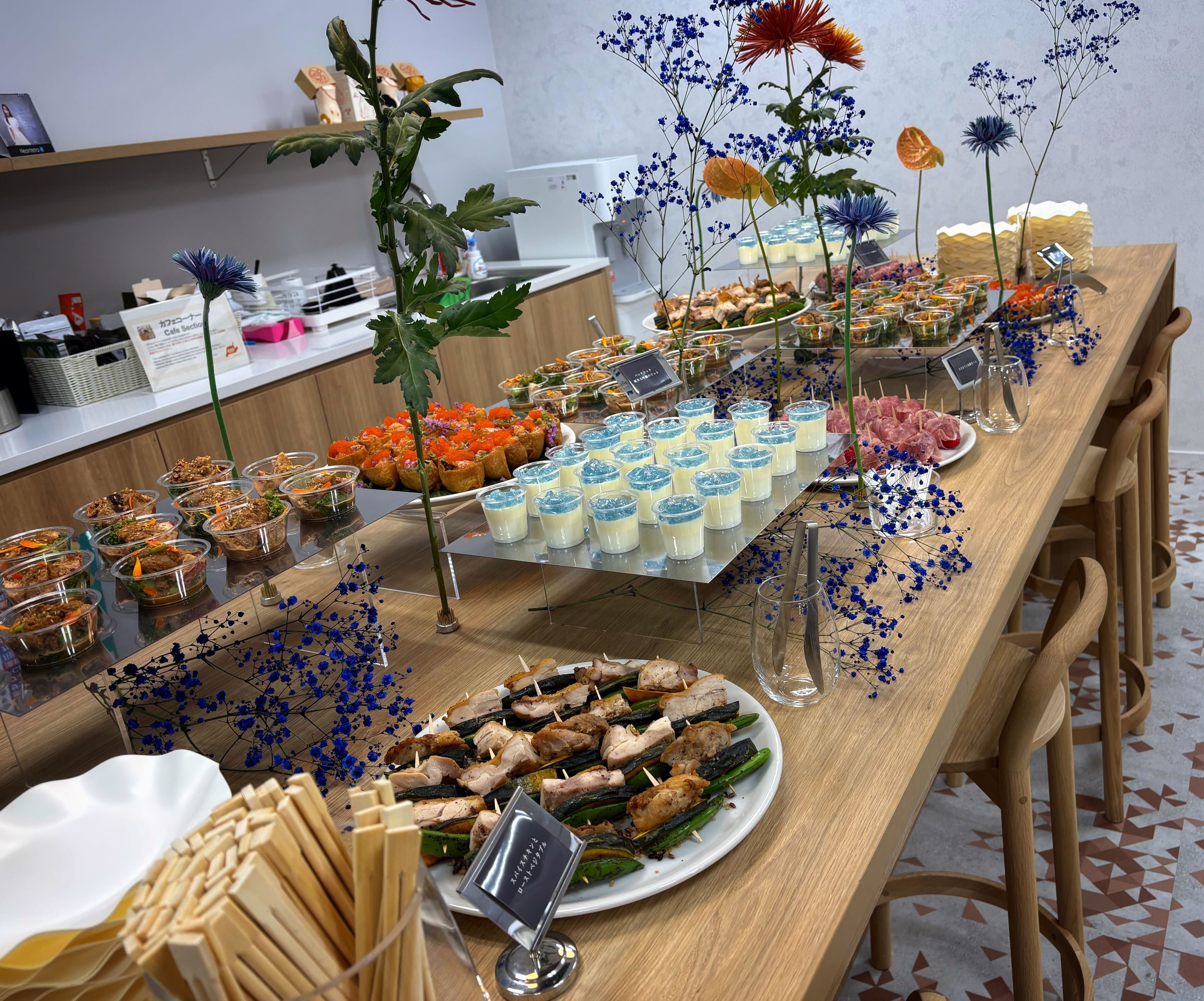 懇親会のケータリング。木目のハイテーブルに、串料理やフィンガーフード、デザートのプリンなどが並び、青やオレンジの花が添えられている。