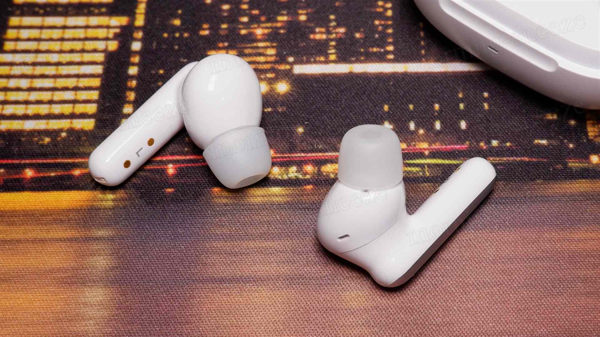 2つの白いワイヤレスイヤフォンがテーブルの上に置かれています。イヤフォンは周囲がぼやけた夜景の背景の上にあり、片方は耳に挿入する部分が透明なシリコン素材で覆われています。もう片方は細い部分がはっきりと見えています。