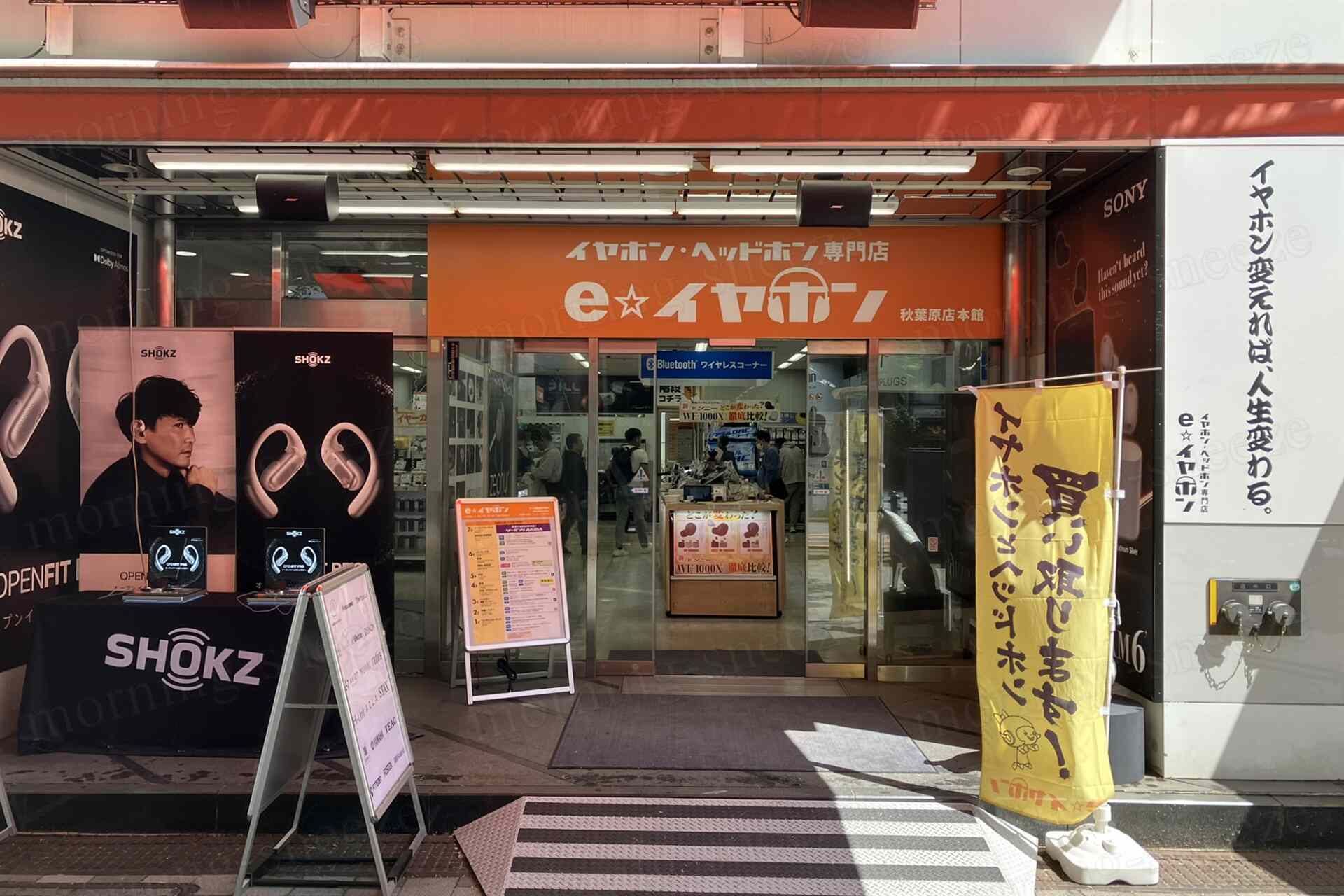 秋葉原にあるイヤホン・ヘッドフォン専門店の外観。店舗の看板はオレンジ色で「e★イヤホン」と表示されている。入り口にはなだらかなスロープがあり、店内が見える。左側にはSHOKZの広告パネルとイヤフォンの展示があり、右側には「イヤホン買い取り中」の旗が立てられている。店の前には案内看板が置かれている。