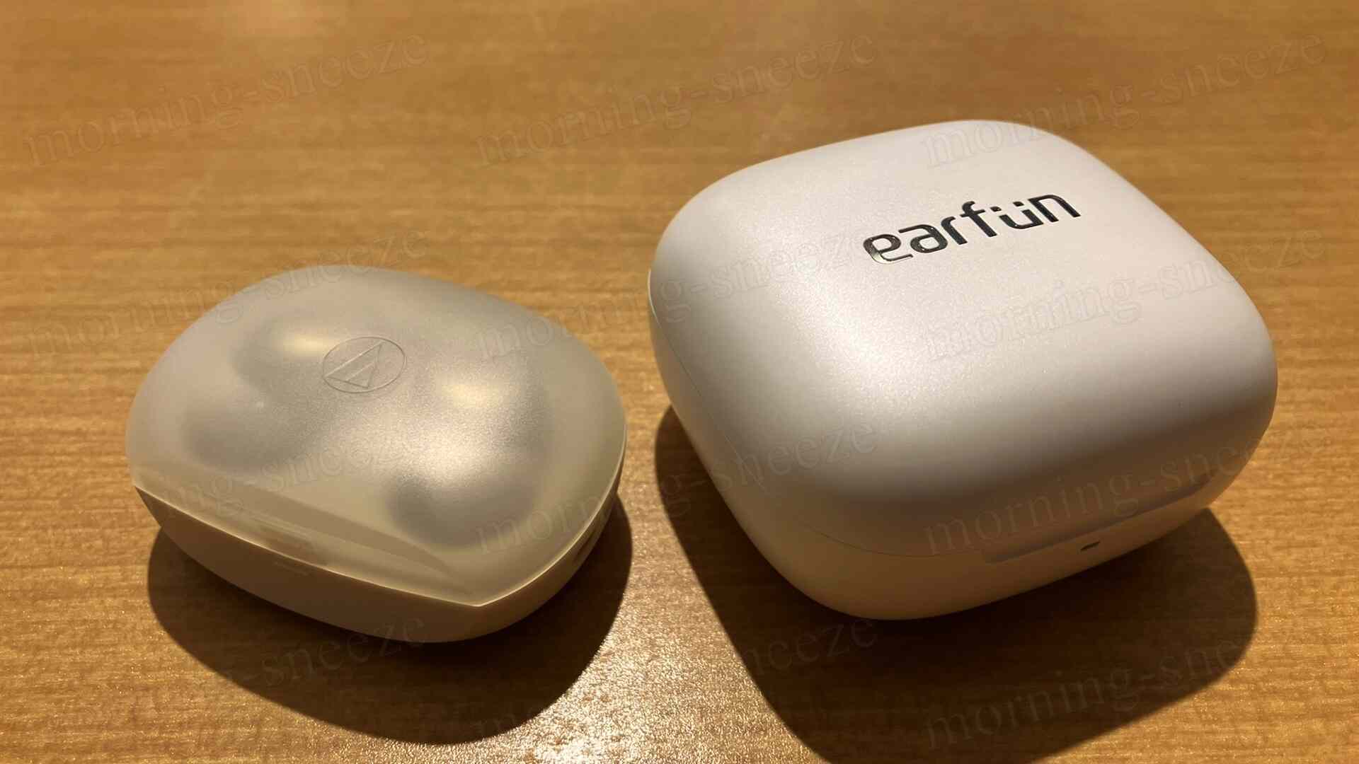 木目調のテーブルの上に置かれた2つのイヤホンケースが写っています。左側には少し透明なケースがあり、その中にはイヤホンが収納されています。右側には「earfun」というロゴが刻まれた白いケースがあります。