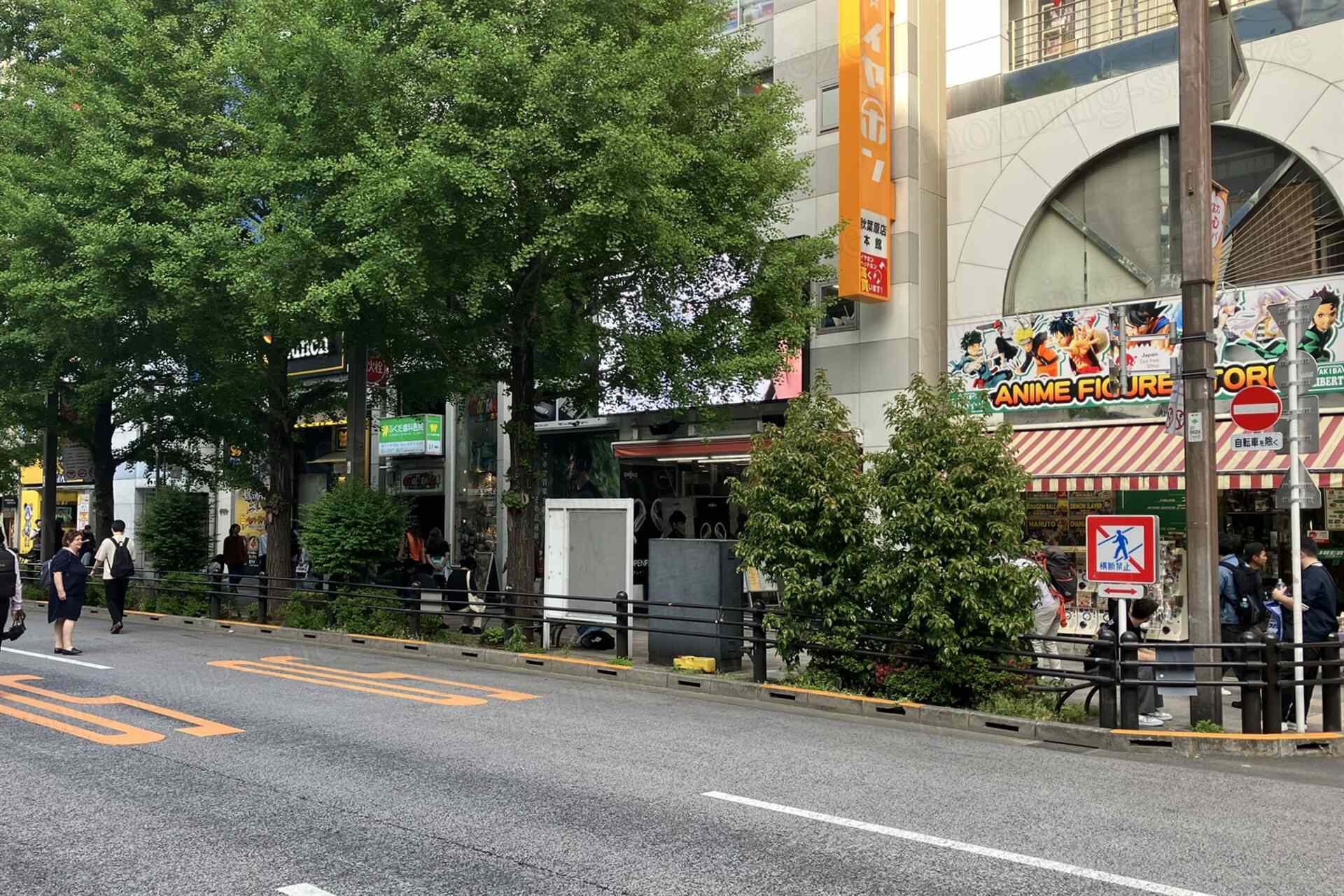 秋葉原電気街通り。赤と白のストライプの看板があるアニメフィギュアショップが並ぶ通り。歩道には緑の木々が生い茂り、数人の人々が通行している。道路には歩行者通行禁止の標識が立てられ、周囲のビルの外壁には広告が掲示されている。