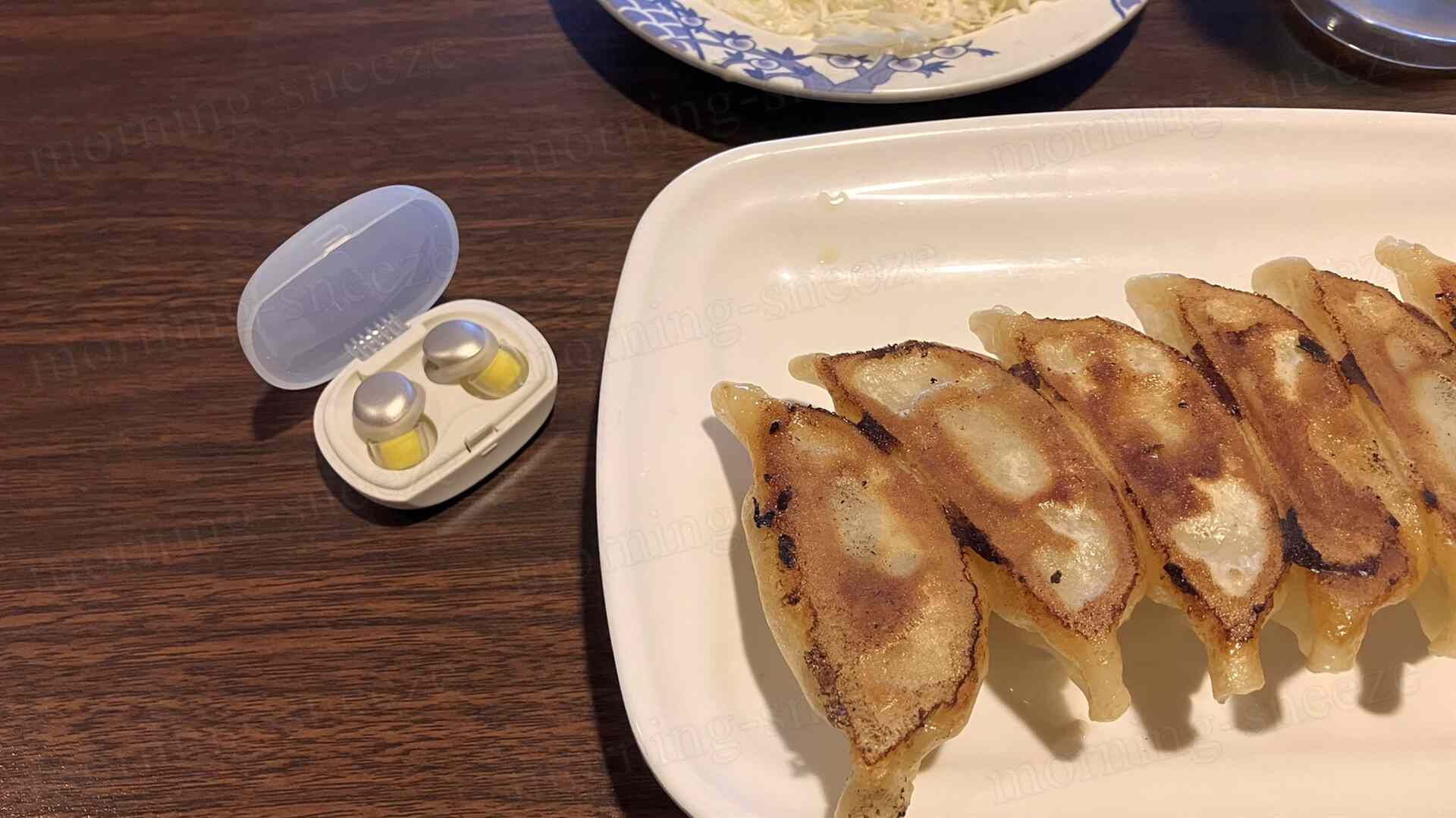 餃子が7つ並んだ白い皿と、その隣に置かれた開いたイヤープラグのケースが見えます。皿の奥にはキャベツの千切りがある皿が見えます。