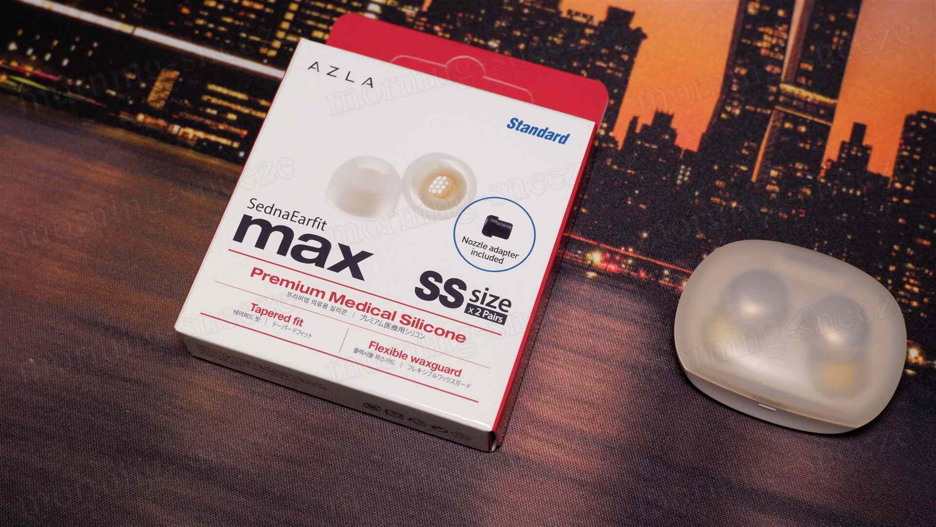 AZLAのSednaEarfit maxのパッケージがテーブルに置かれており、背景には夜景が見える。パッケージは白い箱で、「プレミアム医療用シリコン」と「SSサイズ x 2ペア」の表記がある。また、透明なケースに入ったイヤーピースが隣に見える。