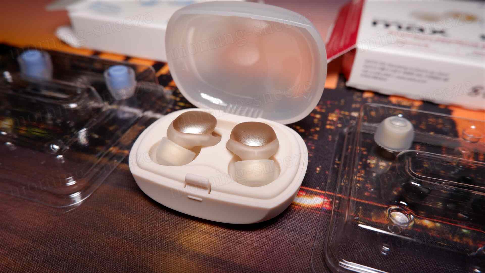 ホワイトのケースに収められたイヤープラグが見える。イヤープラグはクリアで丸みを帯びたデザインで、上部は金色に輝いている。背景には透明なプラスチックのパッケージと、別の部品と思われるものが散らばっている。
