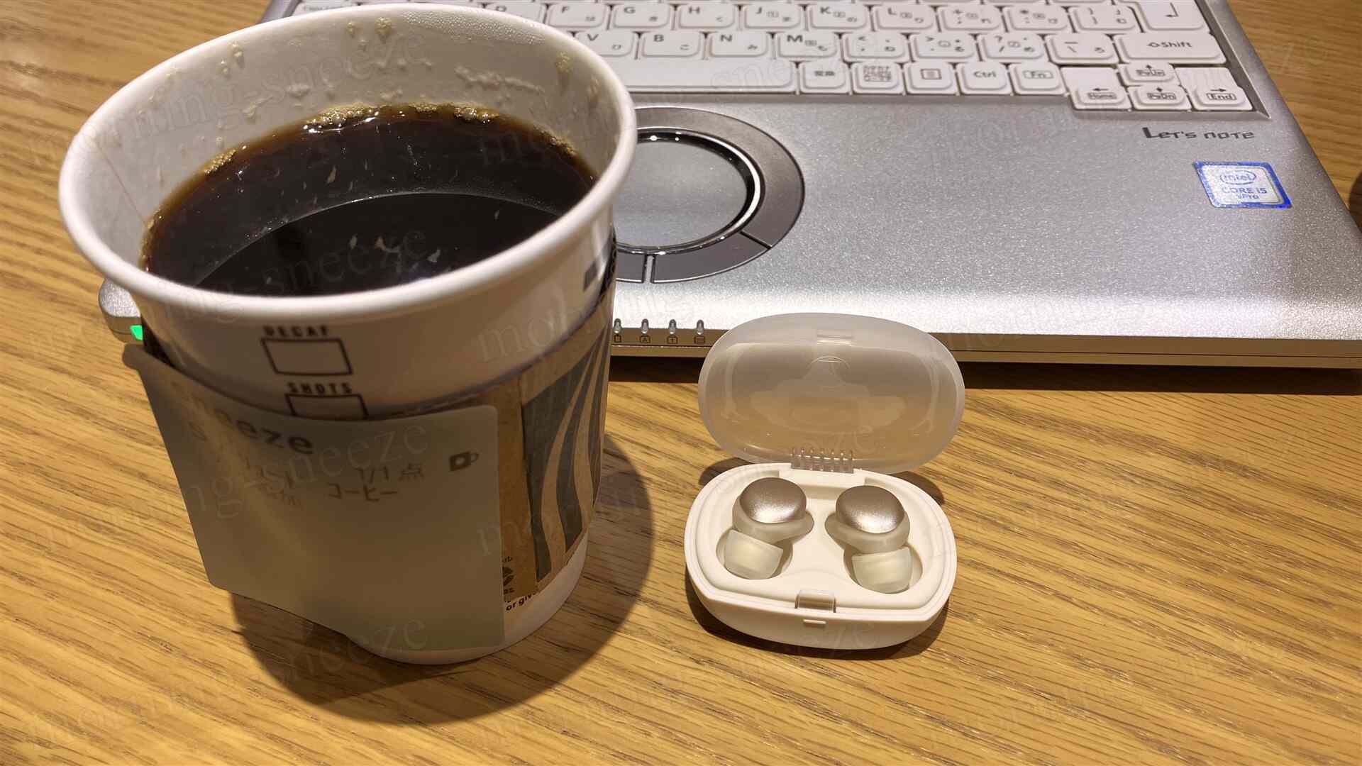 コーヒーの入ったスタバの紙コップ、隣にはイヤープラグが収められたケースがあり、背景にはノートパソコンの一部が見える。テーブルは木製。