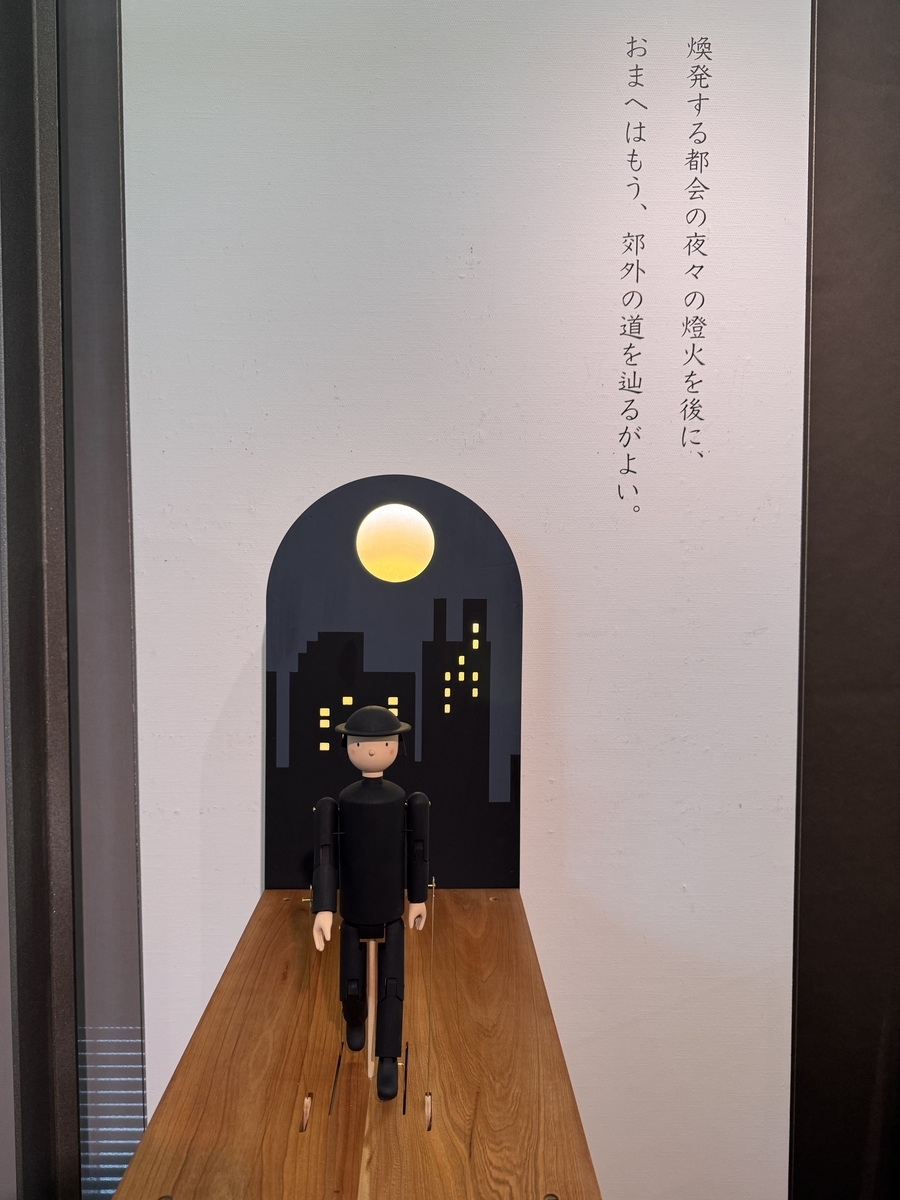 黒ずくめの中也人形が月と東京の街並みの書き割りをバックに歩くオートマタ。背景には「爆発する都会の夜々の燈火を後に、/おまへはもう、郊外への道を辿るがよい。」と中也の詩からの引用が書いてあります。