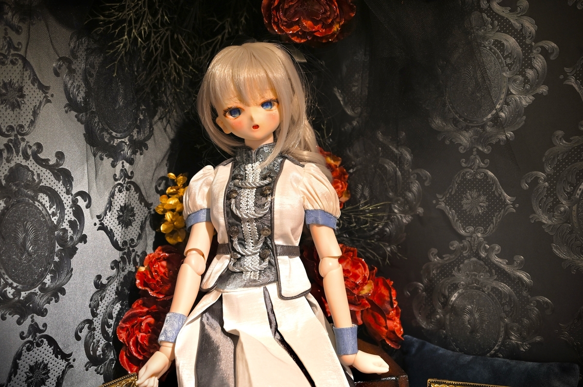黒い壁を後ろに座るギンカ。後ろには赤い薔薇の造花。