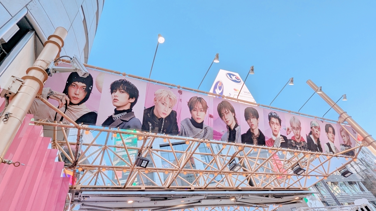 渋谷109の町中に設置された大型広告に、INIメンバー11人のポートレート写真が並ぶ。背景は青空で、UCカードの看板も見える。
