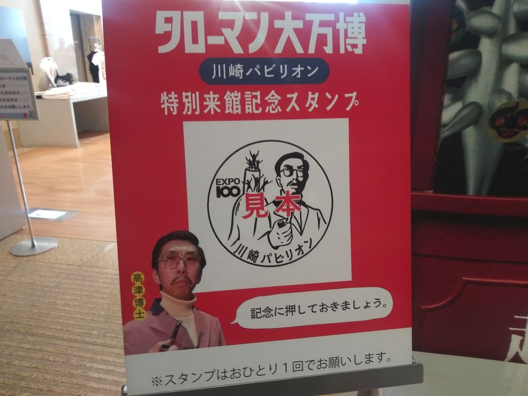 岡本太郎美術館　企画展示室「タローマン大万博川崎パビリオン」入口左のスタンプコーナー　「タローマン大万博川崎パビリオン特別来館記念スタンプ」の案内板　案内板には高津博士が「記念に押しておきましょう。」と言っている吹出付き写真とスタンプ見本。スタンプ見本は「パイプを持つ高津博士の後ろに母の塔」のイラストと「EXPO100」「川崎パビリオン」の文字のあるデザイン。パネル一番下には「スタンプはおひとり1回でお願いします」の注意書きがある