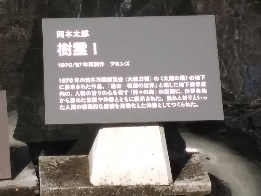 川崎市岡本太郎美術館　階段を上がってすぐ右の池あるに樹霊Ⅰの説明板「岡本太郎　樹霊Ⅰ　1970/97年再制作　ブロンズ　1970年の日本万国博覧会(大阪万博)の＜太陽の塔＞の地下に展示された作品。「過去-根源の世界」と題した地下展示室内の、人間の祈りの心を表す「神々の泉」の空間に、世界から集めた仮面や神像とともに展示された。畏れと祈りといった人間の根源的な勘定を具現化した神像としてつくられたと書かれている
