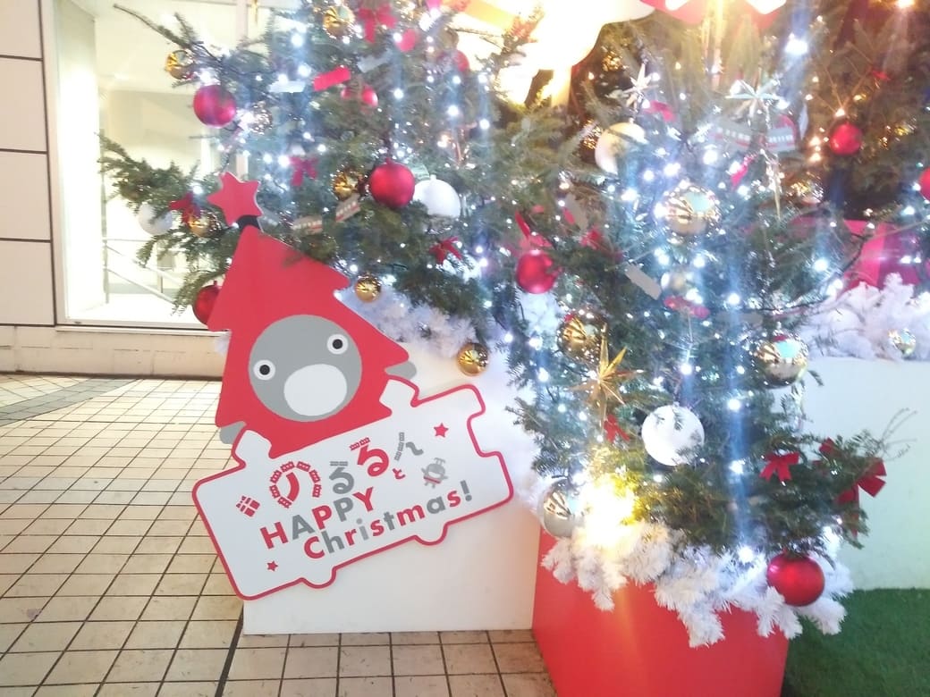 たまプラーザのクリスマスイルミネーション2025年_GATE PLAZA_ステーションコート駅前のクリスマスツリー　左下ののるるんは赤いツリーのかぶり物をしている
