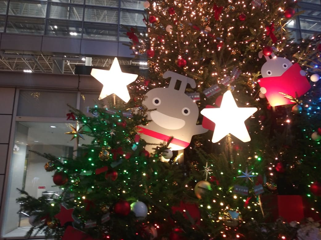 たまプラーザのクリスマスイルミネーション2025年_GATE PLAZA_ステーションコート駅前のクリスマスツリー　えがおののるるんの後ろに本を読むのるるん