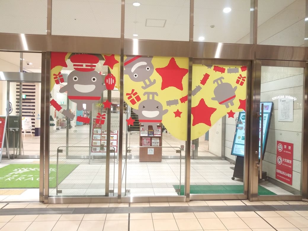 たまプラーザのクリスマスイルミネーション2025年_GATE PLAZA_フェスティバルコート1FモールB口ガラス扉のクリスマスディスプレイは黄色を背景に赤い星と灰色ののるるんが貼られていた