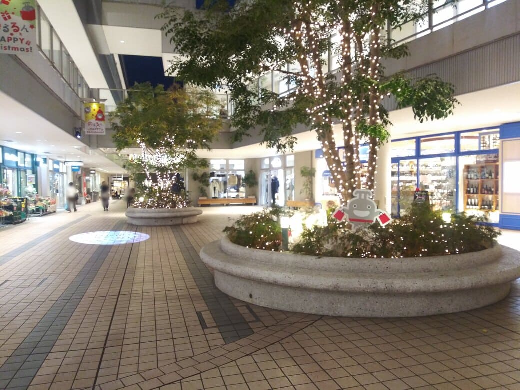 たまプラーザのクリスマスイルミネーション2025年_GATE PLAZA_カルディから交番までの通路　円形にベンチが配された植込みには赤いショップバッグを両手に持ったのるるんが飾られていた