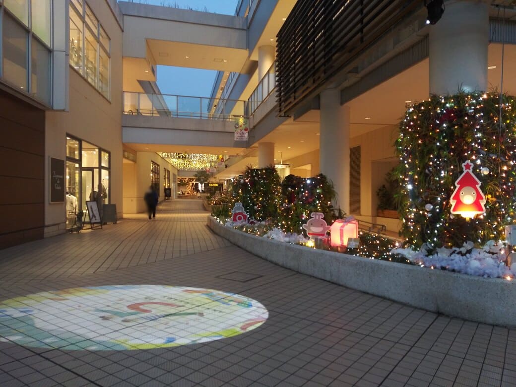 たまプラーザのクリスマスイルミネーション2025年_GATE PLAZA_交番側からカルディ方向への通路　床にはロゴスポットライト、植込みには複数ののるるんの平面オブジェとブレ遷都がライトアップされている