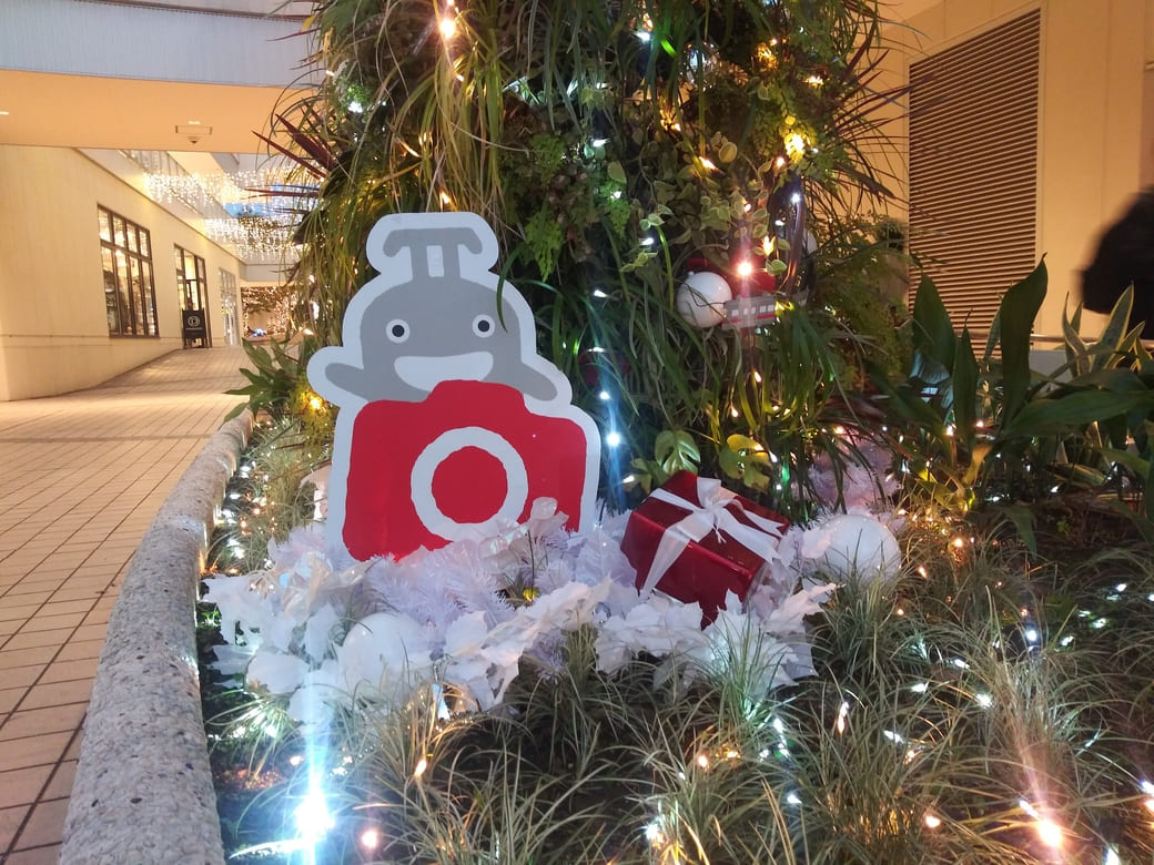 たまプラーザのクリスマスイルミネーション2025年_GATE PLAZA_交番側からカルディ方向への通路植え込みにあるのるるんのオブジェとライトアップされたディスプレイ　プレゼントの包みと笑顔ののるるんの後ろには赤いカメラを抱えたのるるんのオブジェ