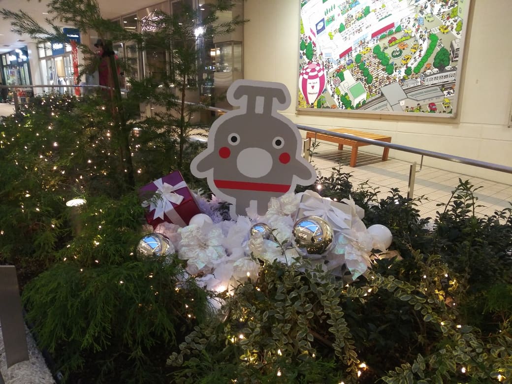 たまプラーザのクリスマスイルミネーション2025年_GATE PLAZA_交番側からカルディ方向への通路植え込みにあるのるるんのオブジェとライトアップされたディスプレイ　のるるんは丸い口を大きく開けて立っているその手前には白いリボンで結ばれたえんじ色のプレゼントの包みと白いプレセントの包み、銀入りのボールのオブジェと小さな暖色系のライトが連なって灯っている