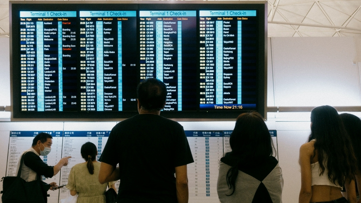 空港の大型電光掲示板で出発便のチェックイン状況を確認する、マスク姿の人々の後ろ姿。