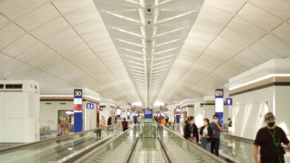 香港国際空港の広々とした出発ロビー。高い幾何学的な天井の下、多くの利用客が動く歩道で行き交う様子。