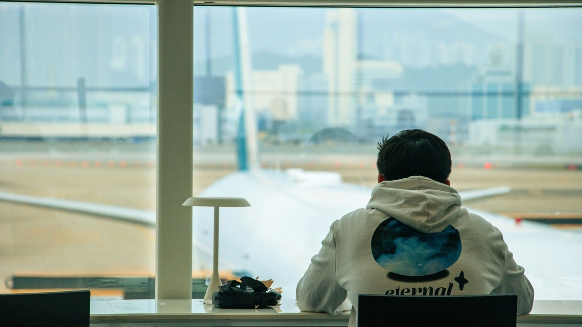 空港の待合エリアで、窓の外に駐機しているキャセイパシフィック航空の機体を静かに眺める白いパーカーの男性。