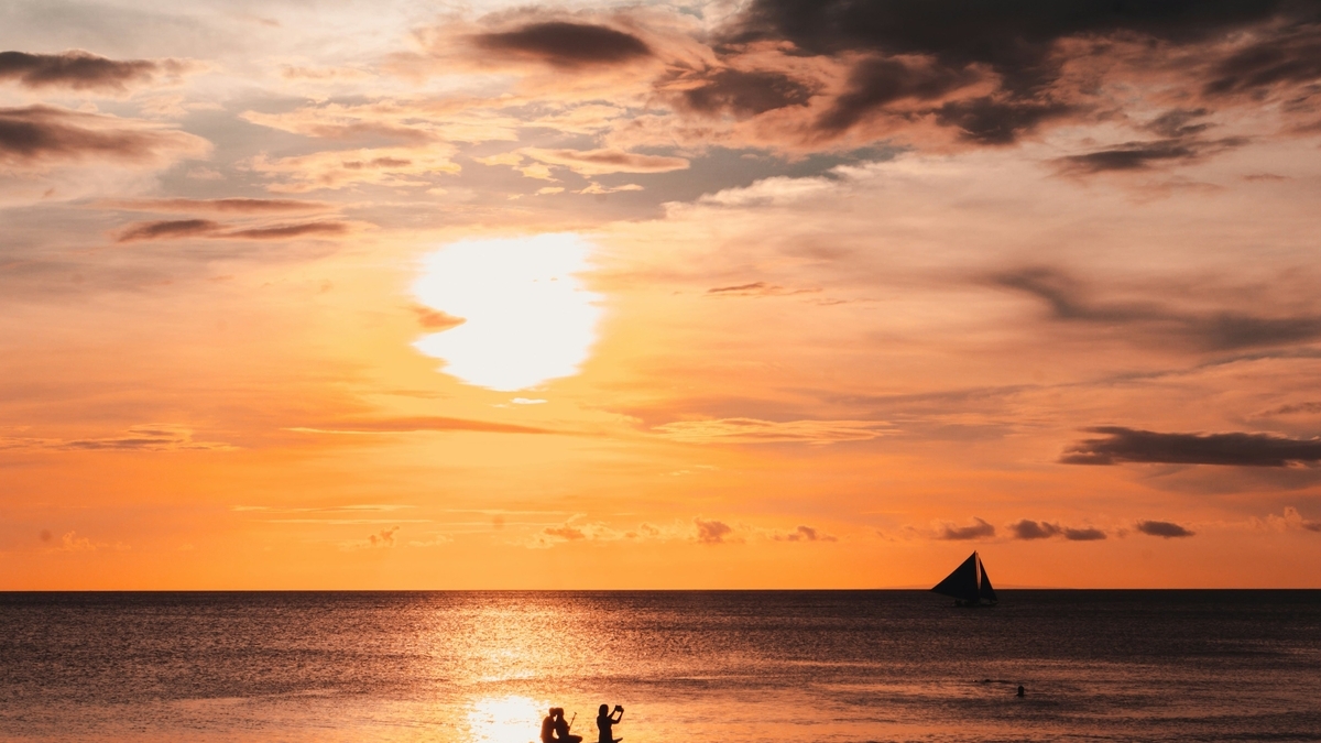 オレンジ色の夕日に照らされた海辺で、一艘のヨットが波間に浮かび、手前の砂浜で数人の人々が夕景を眺めたり撮影したりしている様子