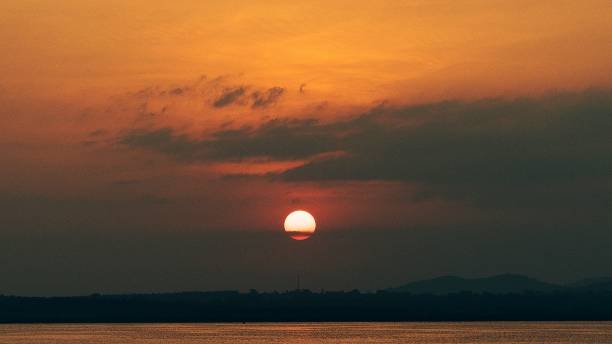 山並みのシルエットが遠くに見える水辺で、厚い雲の隙間から丸く赤みを帯びた夕日が沈み、空全体が深いオレンジ色に染まる風景