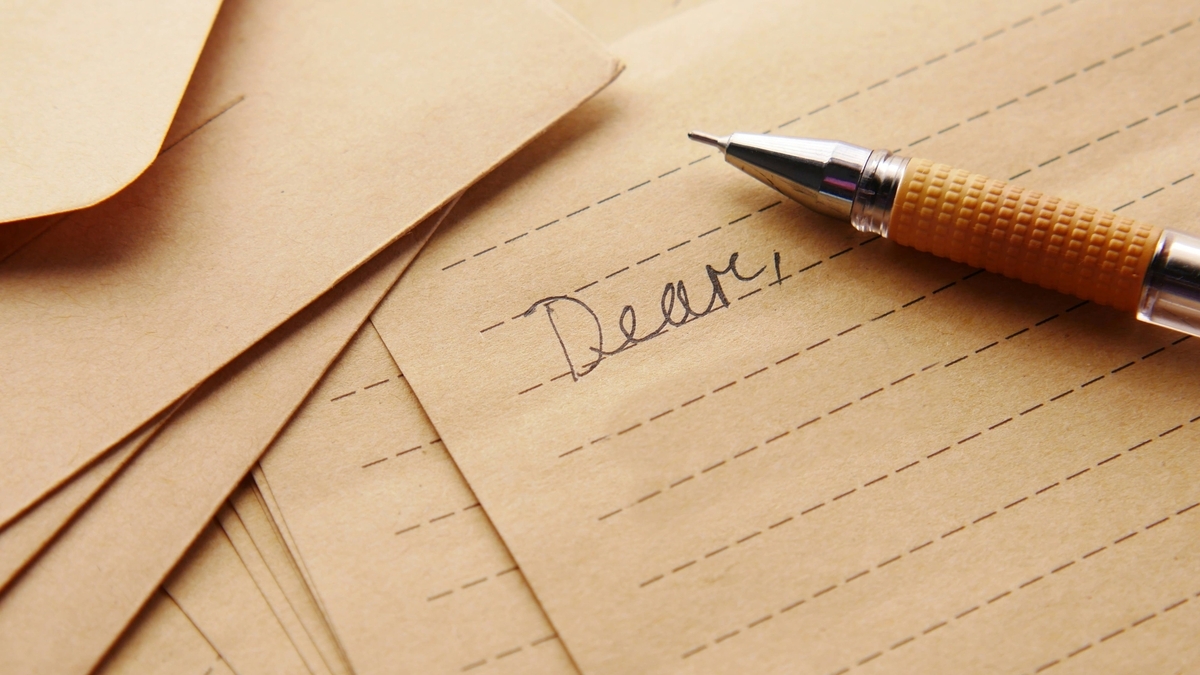 罫線が引かれた茶色の便箋に、黒いペンで「Dear,」と手書きで書き始められており、横にはオレンジ色のグリップのシャープペンシルが置かれている様子