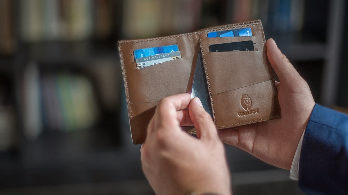 茶色のレザー製二つ折り財布を両手で持ち、中に入っている複数のクレジットカードやビジネスカードを確認している様子。背景にはぼけた本棚が写り込んでいる