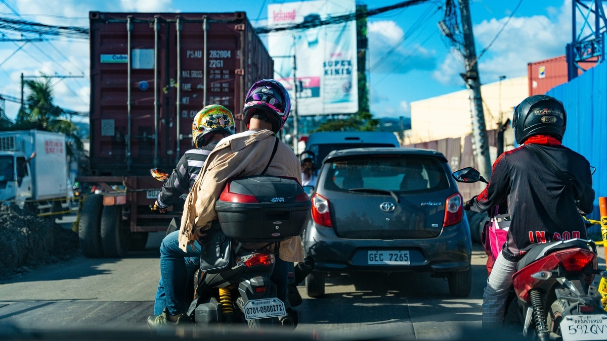 ヘルメットを被り荷物を積んだ複数のバイクや乗用車、大きなコンテナを載せたトラックが、電線の張り巡らされた青空の下で混雑する道路を走行している後方からの風景