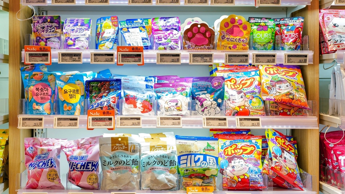 日本のコンビニやスーパーの棚に、ハイチュウやミルクキャンディなどの色とりどりの菓子パッケージが陳列されている様子。各商品の前には価格やセール情報が記載された値札がついている