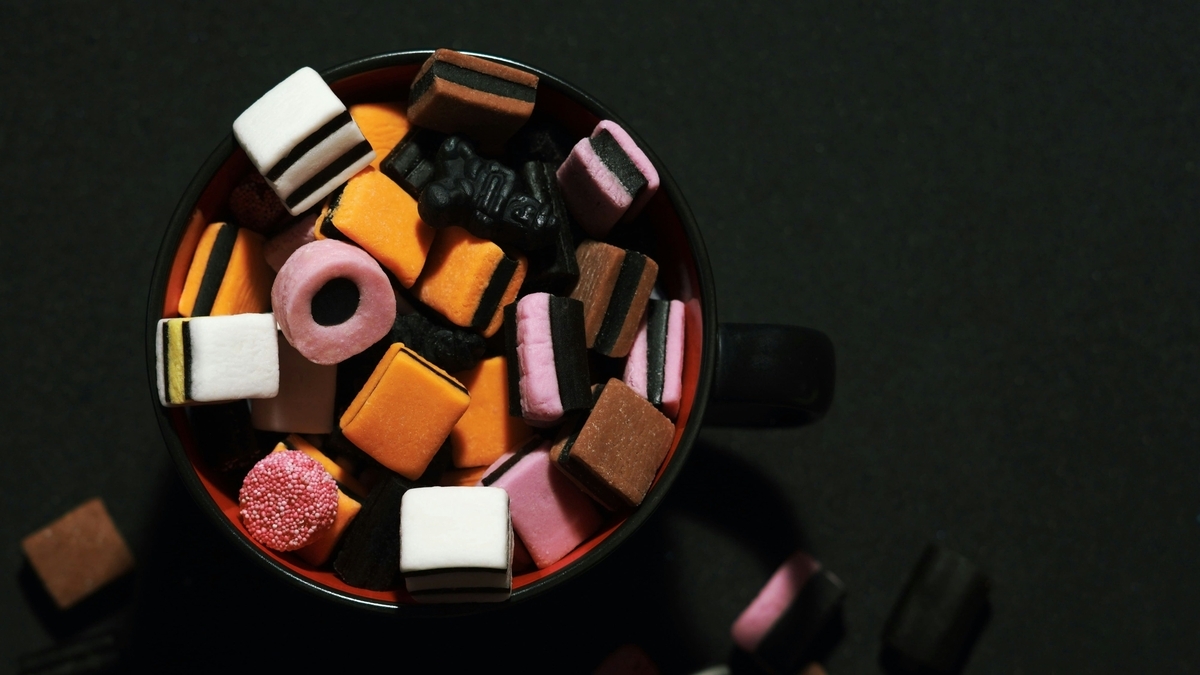 マグカップの中に、黒、オレンジ、白、ピンクなど色鮮やかなリコリス菓子やアソートキャンディが山盛りに詰められている様子。真上から撮影されており、背景は黒く統一されている。