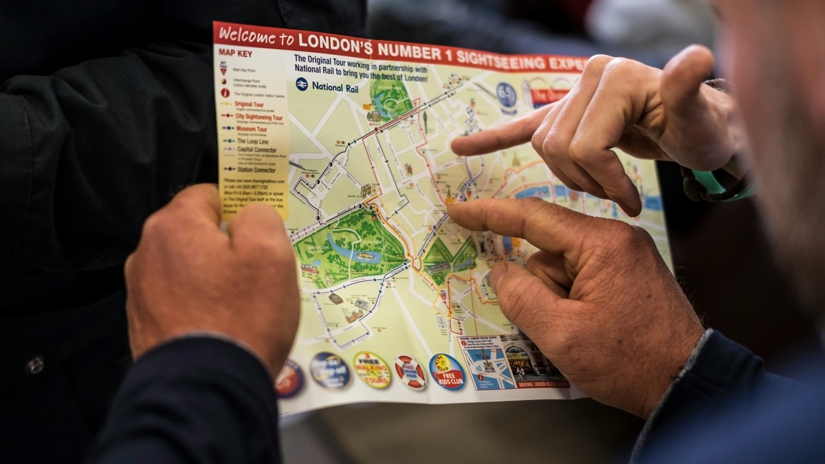 「Welcome to LONDON」と書かれた観光地図を、二人の人物が指で指し示しながら確認している様子。地図にはロンドンの主要な通りや鉄道路線が細かく描かれており、指先は特定の地点を指している。
