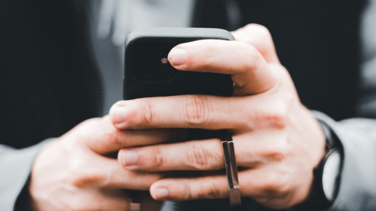黒いケースに入ったスマートフォンを両手で持ち、操作している人物の手元のアップ。指先が画面に触れており、指には金属製のリングがはめられている。背景はボケており、スマートフォンの質感に焦点が合っている。
