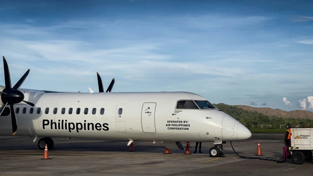 青空の下、滑走路に停車しているフィリピン航空の白いプロペラ機。機体には「Philippines」のロゴがあり、前方では地上スタッフが作業を行っている。背景には緑豊かな山々が見える。